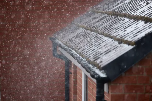 Heavy rain on roof This architecture photograph captures heavy rain falling on the roof of a residential brick house during the afternoon in early autumn. The rain is visibly streaming across the roof tiles and down the gutters, emphasizing the intensity of the downpour. The image highlights the urban setting, with the brick walls and window of the house clearly visible. The afternoon light is diffused by the dense rain, which is characteristic of autumn weather, adding a dynamic and atmospheric quality to the scene.